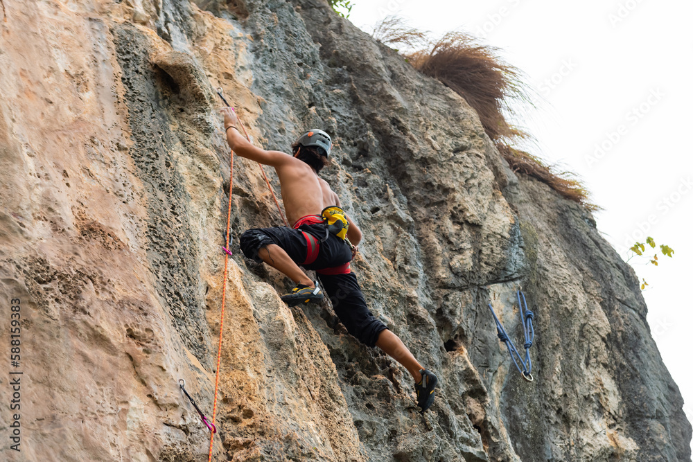 Young Asian man climber climbing on rocky coastline at tropical island in sunny day. Handsome guy enjoy outdoor active lifestyle and extreme sport training mountain climbing on summer holiday vacation