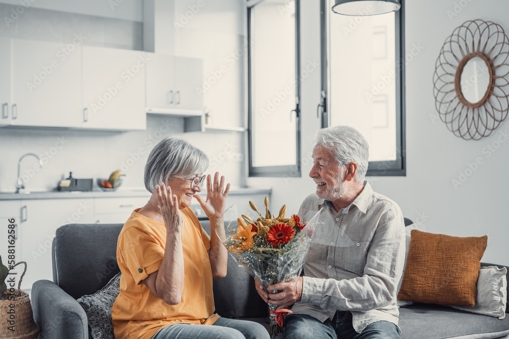 Old man giving flowers at his wife sitting on the sofa at home for the San Valentines’ day. Pensioners enjoying surprise together. In love people having fun.. Old man giving flowers at his wife sitting on the sofa at home for the San Valentines’ day. Pensioners enjoying surprise together. In love people having fun..