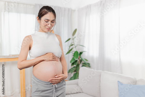Free Download pregnant Asian people stand in white room, she use her hand touch on her abdomen, she feeling love and care her daughter, pregnancy health care promotion - GFXLoad