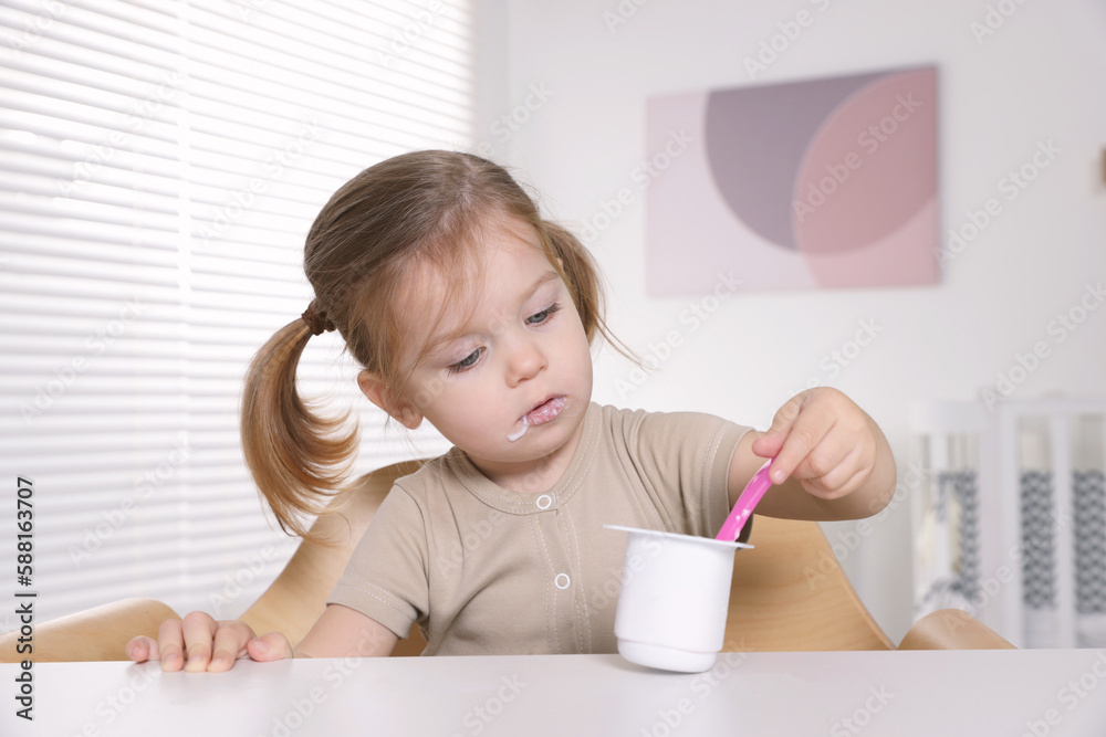 Cute little child eating tasty yogurt from plastic cup with spoon at white table indoors Cute little child eating tasty yogurt from plastic cup with spoon at white table indoors