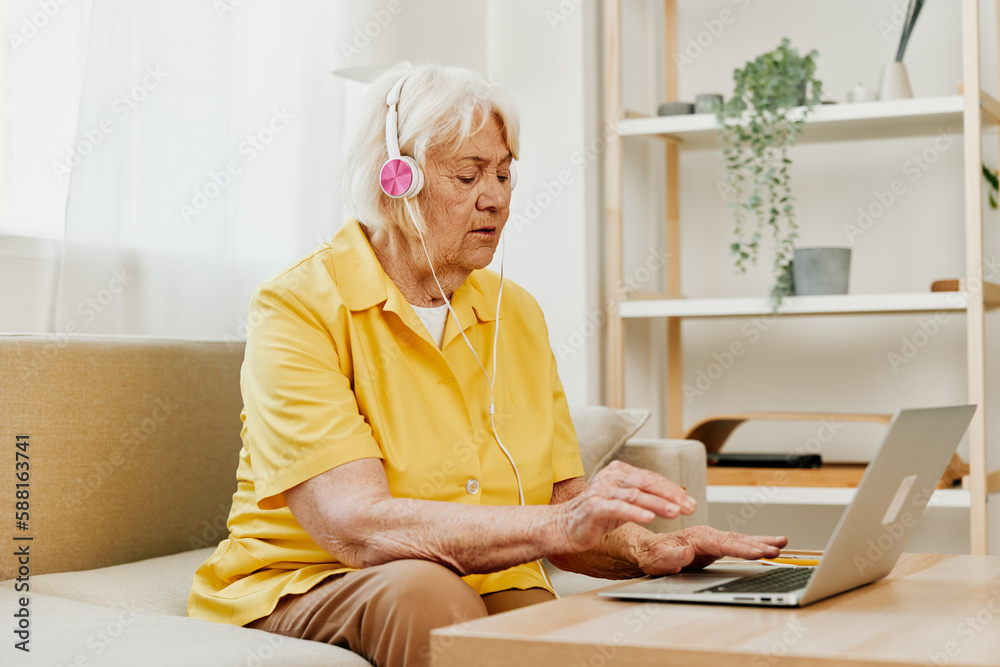 An elderly woman wearing headphones with a laptop sits on the couch at home and works in a yellow shirt in front of a window. An elderly woman wearing headphones with a laptop sits on the couch at home and works in a yellow shirt in front of a window.