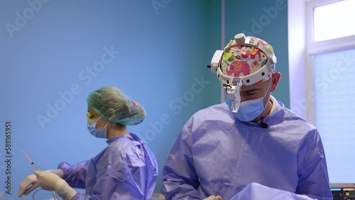 Calm serious surgeon performs operation in surgery room. Nurse prepares tools at backdrop. photo