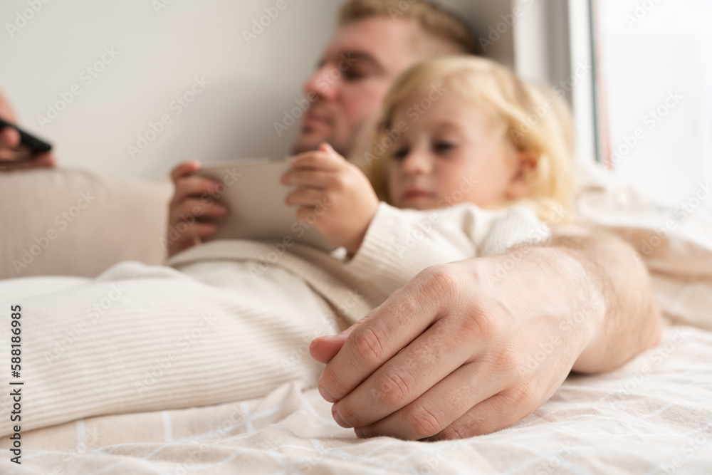 Defocused girl and adult man lying on bed using phones at home. Child and father looking at gadgets, spending time together.