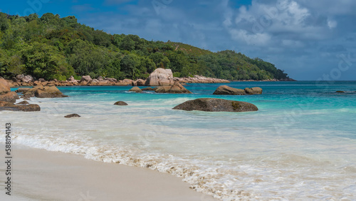 The waves of the turquoise ocean foam on the white sand of the beach. Picturesque boulders in the surf. A hill overgrown with tropical vegetation against a blue sky and clouds. Seychelles. Praslin.