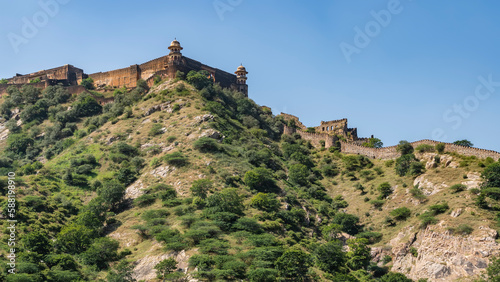 The ancient fortress wall runs along the ridge of the mountain. Watchtowers with domes are visible. Blue sky. Green vegetation on the slope. India. Amber Fort. Jaipur