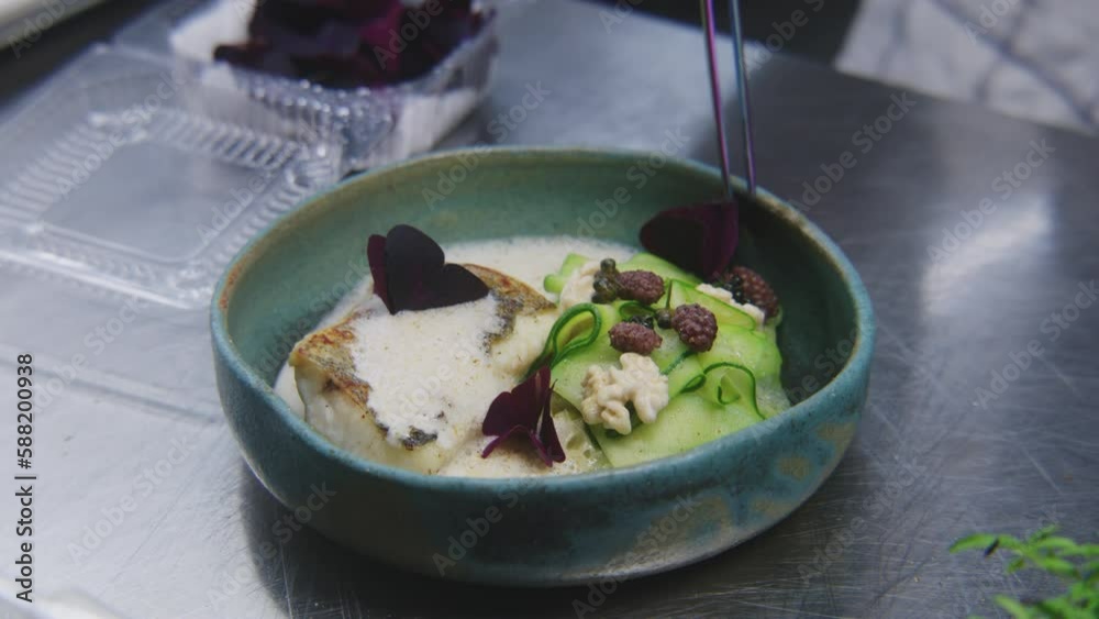 Male cook makes presentation of dish. The chef decorates dish of fried fish and stewed vegetables in bechamel sauce with flower petals on top. Restaurant with professional gourmet cuisine. Close up.