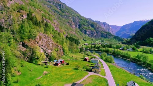 Aerial drone backward moving shot over a river flowing along the valley surrounded by mountain range in Norway on a sunny day. Village houses along roadside. photo