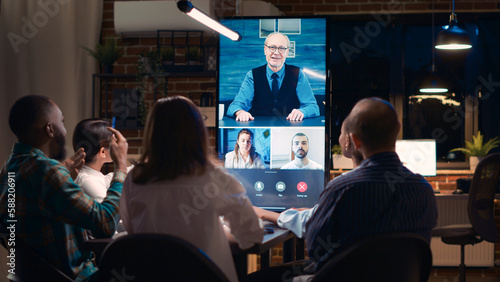 Employees greeting elderly executive in videocall, waving hi, colleagues videoconference communication. Office workers listening to team lead speaking, talking in teleconference at night. Tripod shot.