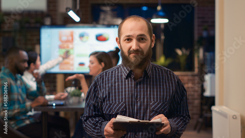Employee reading report, holding clipboard, looking at camera, smiling. Young man medium shot portrait, coworkers talking in business meeting statistics presentation in background. Handheld shot.