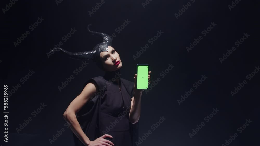 Woman in witch costume for costume party stands with smartphone with green screen in hand, black background, indoor, front view. . Concept of cosplay for halloween.