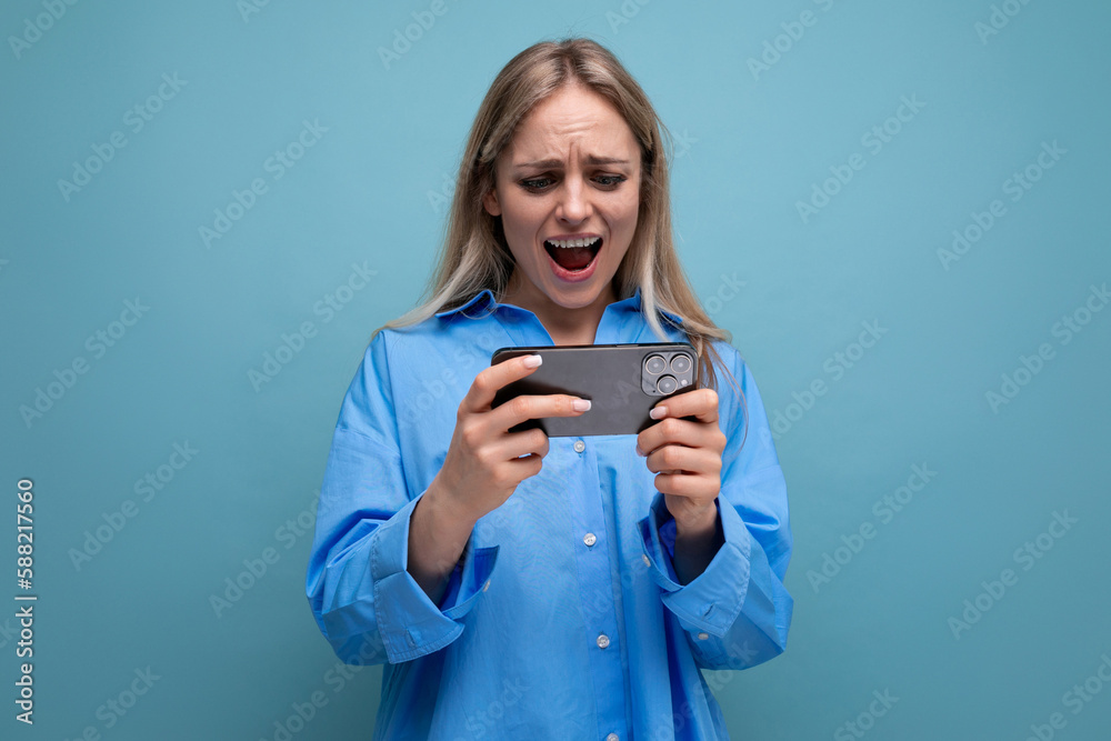 joyful charming blonde girl shocked holding smartphone in hands over isolated blue background joyful charming blonde girl shocked holding smartphone in hands over isolated blue background