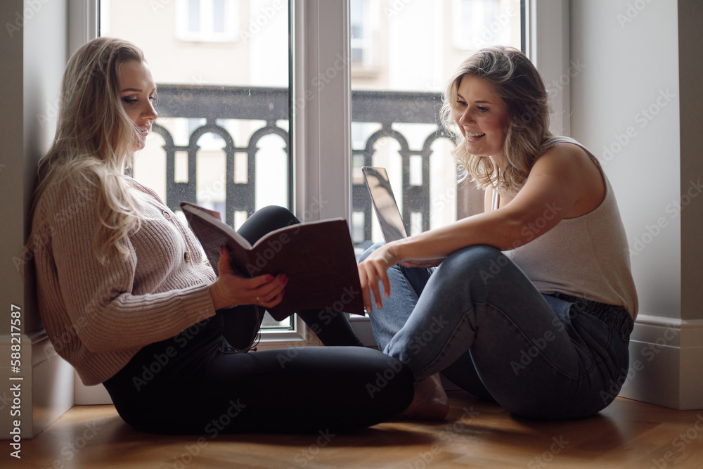 Happy beautiful women sitting together on floor near access to balcony, talk and read book. Home leisure, female talk. Happy beautiful women sitting together on floor near access to balcony, talk and read book. Home leisure, female talk.