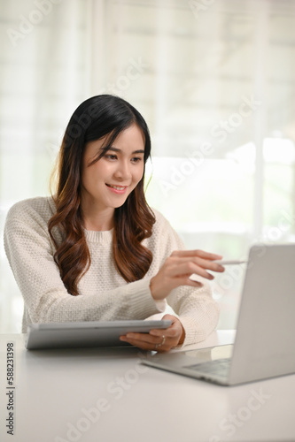 A beautiful Asian woman pointing pen at a laptop screen, working in her home office.