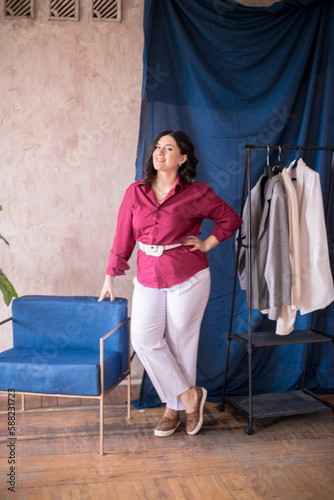 A young stylist girl with dark hair smiles in a trendy burgundy white jacket and light trousers in a light studio with green live plants in pots and a rack with clothes