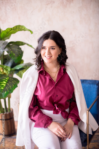 A young girl stylist with dark hair smiles in a trendy white jacket, a burgundy color shirt and light trousers in a light studio with green living plants in pots  © capable97