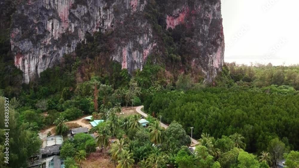 Aerial view of massive tree covered mountain with steep cliff, tropical jungle forest and houses around in Ko Libog, Mueang Trang, Thailand. Idyllic evening golden dusk scenery, drone flying downward Aerial view of massive tree covered mountain with steep cliff, tropical jungle forest and houses around in Ko Libog, Mueang Trang, Thailand. Idyllic evening golden dusk scenery, drone flying downward