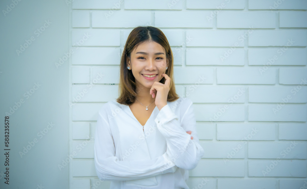 Beautiful asian business woman is standing with her hand on her cheek and smiling with a white brick wall in the background, Digital marketing. Beautiful asian business woman is standing with her hand on her cheek and smiling with a white brick wall in the background, Digital marketing.