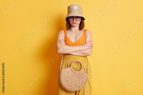 Indoor shot of offended sad young woman wearing summer clothing with straw bag standing with folded arms and pout lips isolated over yellow background
