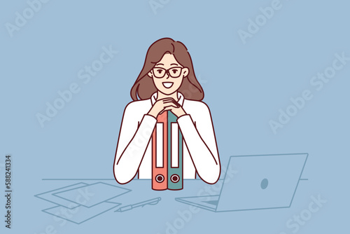 Business woman sits at table leaning on folders and smiling looking at camera rejoicing at success in career as office clerk. Girl manager with documents on desk rejoices at end of business audit