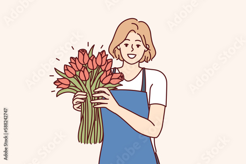 Woman flower seller stands with luxurious bouquet and smiling looks at camera enjoying work of florist in flower shop. Positive girl in apron holds fragrant bouquet with scarlet buds 