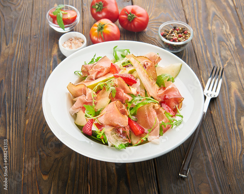 Delicious summer salad in white plate with sweet pears, strawberry, jamon and fresh arugula on wooden background. Healthy organic eating concept