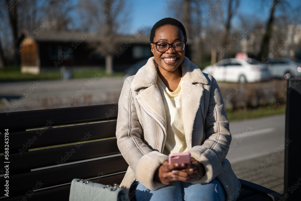 Obraz premium Outdoor portrait of happy african-american woman on sunny day. She is sitting on bench in the street and messaging on smartphone.