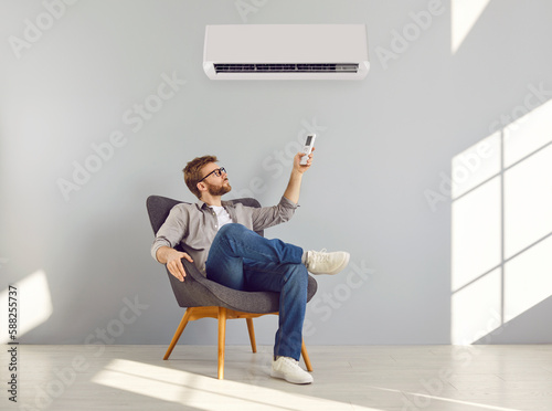 Young bearded man in glasses and casual clothes turning on air conditioner sitting on chair in a empty space enjoying cool conditioned air using remote isolated on a grey wall background.