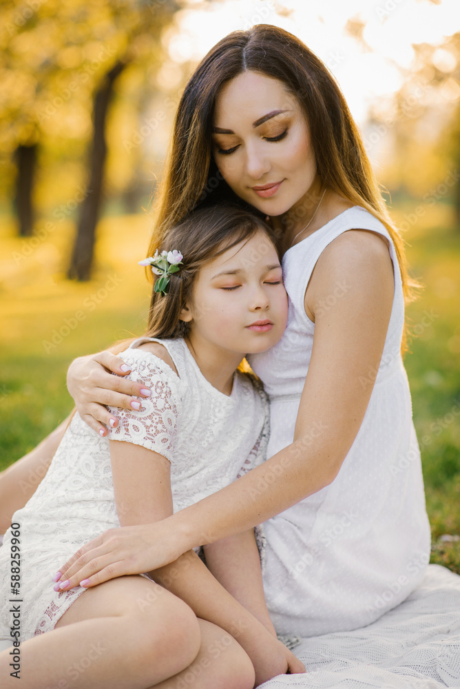 A happy young woman sits on a picnic blanket and hugs her daughter. mother and daughter's summer picnic in the apple orchard. A happy young woman sits on a picnic blanket and hugs her daughter. mother and daughter's summer picnic in the apple orchard.