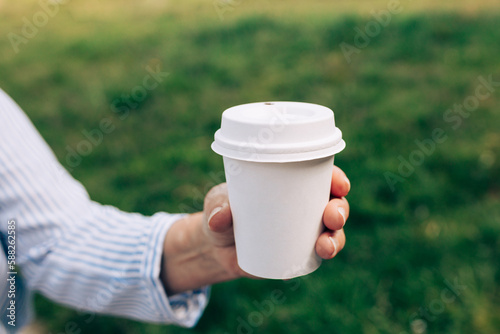Womans hand holding white paper cup of coffee on natural morning background. Place for logo. Coffee to go. Disposable paper cup closeup. Delicious hot beverage. Blank space for text, mockup