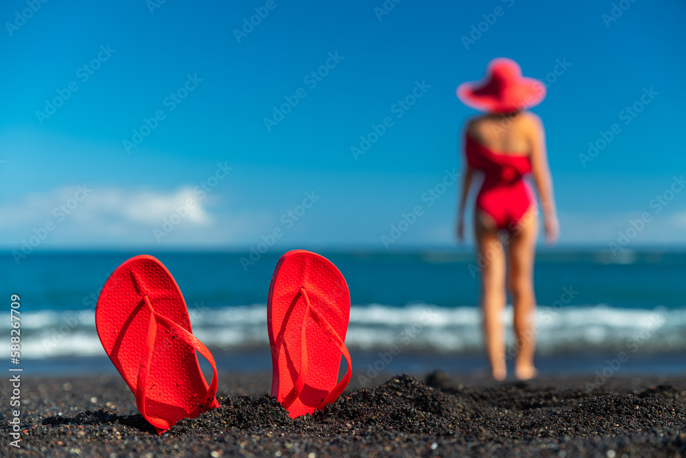 Red flip flops in the black sand and silhouette of woman in red swimsuit standing on the ocean beach. Summer concept. Red flip flops in the black sand and silhouette of woman in red swimsuit standing on the ocean beach. Summer concept.