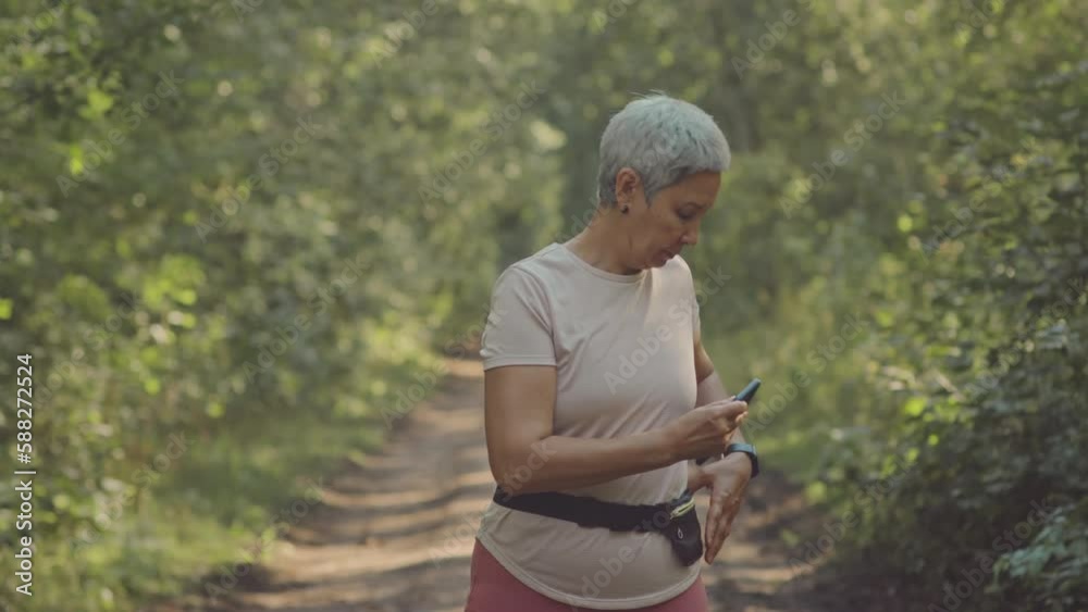Medium shot of mature Asian woman taking her smartphone out of waist bag while jogging outdoors in park on sunny day Medium shot of mature Asian woman taking her smartphone out of waist bag while jogging outdoors in park on sunny day