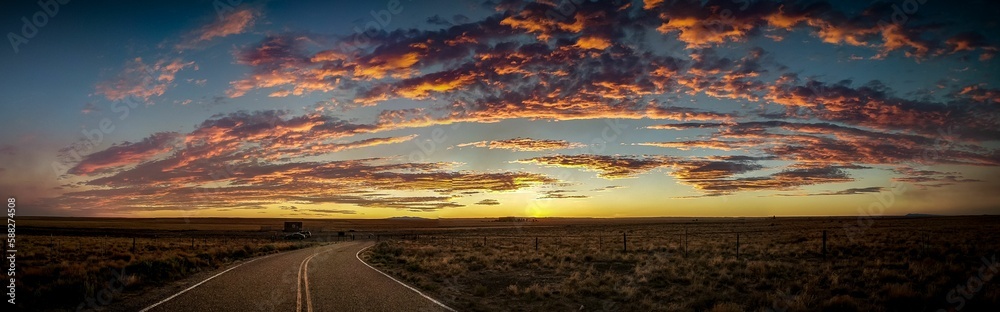 Panoramic shot of a sunrise with purple clouds and blue sky, asphalt and fields on both sides
