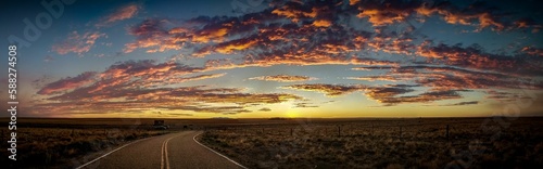 Panoramic shot of a sunrise with purple clouds and blue sky, asphalt and fields on both sides