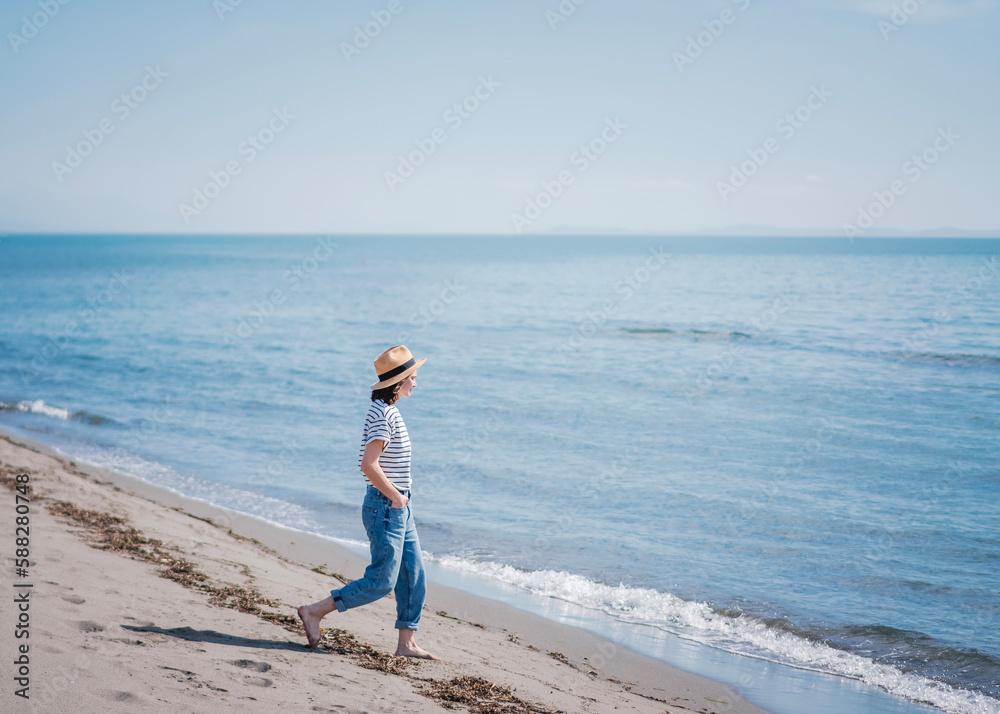 Young happy woman in a hat walking along the sandy beach on the blue sea. Summer holidays and travel concept