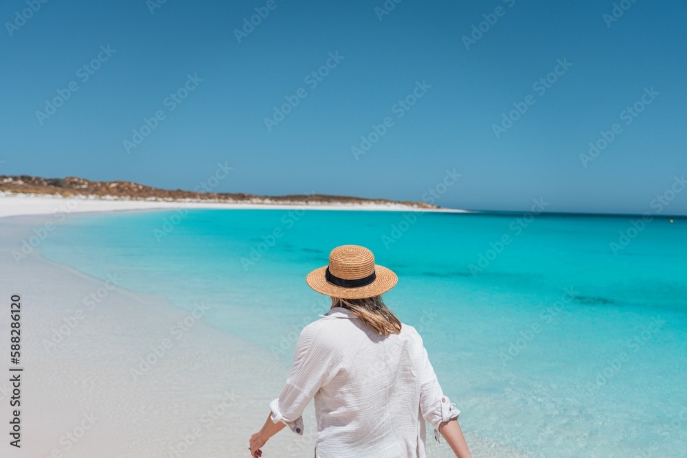 Caucasian female with a straw hat walking on a sandy beach along blue sea on a sunny day