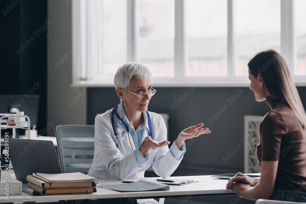 Obraz premium Senior female doctor talking to patient and gesturing while sitting at the medical office