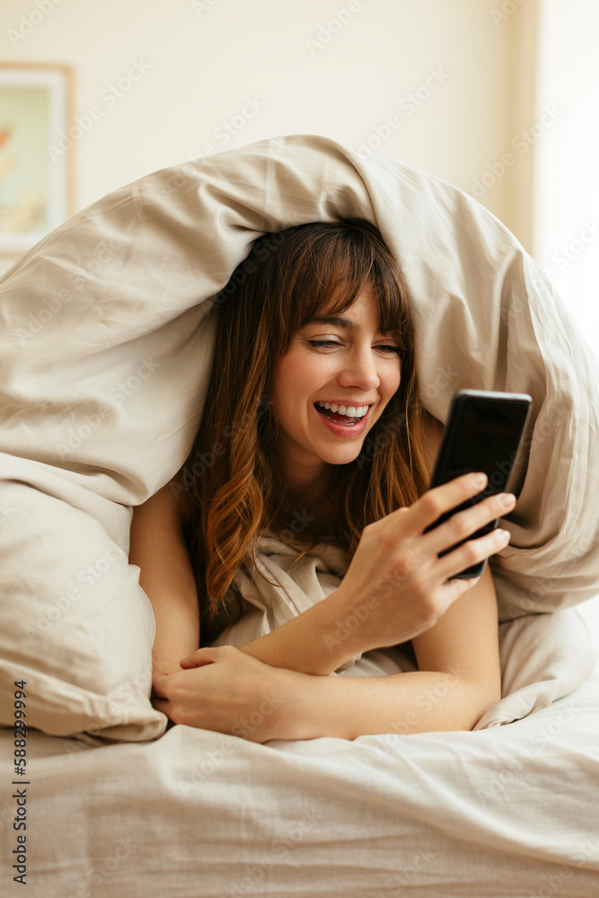 Happy woman using smart phone wrapped in blanket at home Happy woman using smart phone wrapped in blanket at home