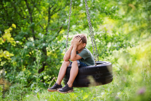 sad boy covering his face with his hands in a swing tire in the forest. Quarrel with friends, resentment, loneliness