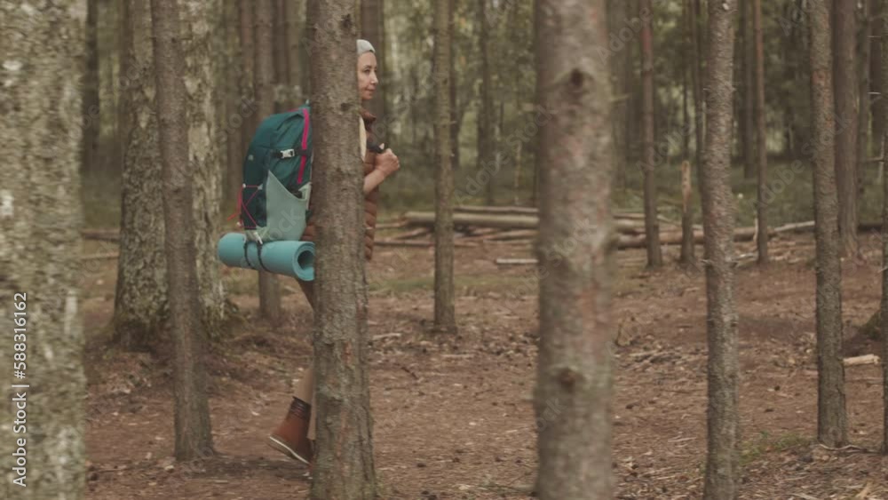 Tracking side view full length shot of active adult Asian woman in wireless headphones with rucksack on her back hiking on footpath in forest Tracking side view full length shot of active adult Asian woman in wireless headphones with rucksack on her back hiking on footpath in forest