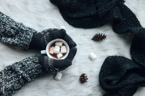 Hands using warm winter clothes and gloves hold a cup oh hot chocolate marshmallow on white fur rug with black knitted scarf. Top view.