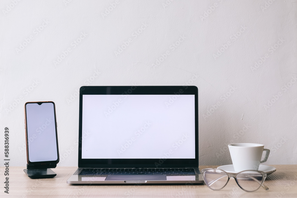 White blank screen laptop computer and smartphone with coffee and eyeglasses on work table at minimalist home office. Front view and mock up. White blank screen laptop computer and smartphone with coffee and eyeglasses on work table at minimalist home office. Front view and mock up.