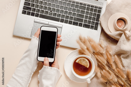 Woman hands hold mobile phone at laptop background with tea, candle, lagurus grass on linen cloth, aesthetic home workplace, cozy feminine office desk. Online shopping, work and business branding