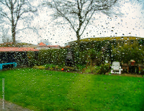 Durch eine Fensterscheibe mit Regentropfen im Frühling bei Schlechtwetter sieht man einen frischen grünen Garten