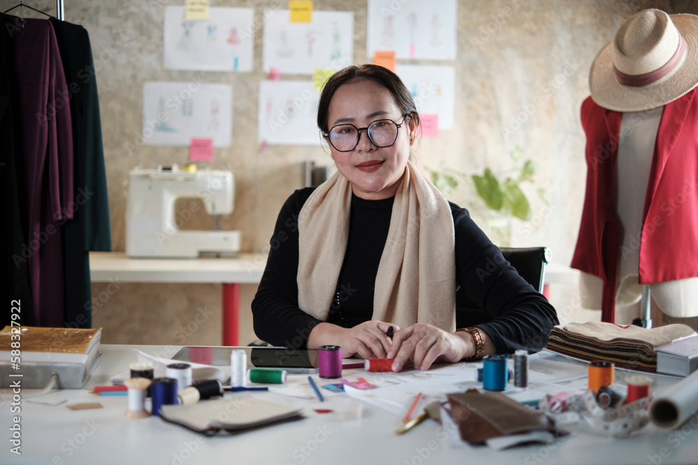 Asian mature female fashion designer is sitting at table, looking at camera and in studio, working with creative and sewing for dress design collection, professional boutique tailor SME entrepreneur. Asian mature female fashion designer is sitting at table, looking at camera and in studio, working with creative and sewing for dress design collection, professional boutique tailor SME entrepreneur.