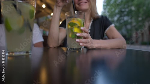 Close-up healthful drink in glass with happy smiling unrecognizable woman stirring cocktail. Relaxed Caucasian lady sitting in cafe with juice enjoying leisure on vacations