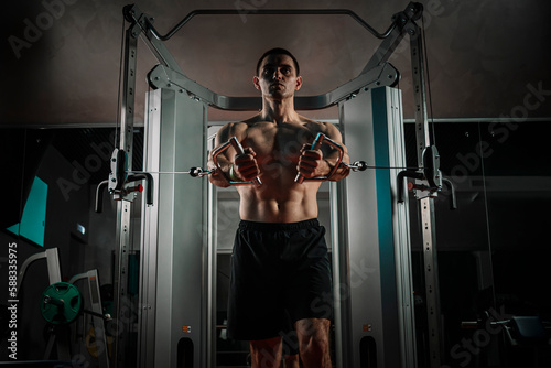 A bodybuilder with a beard is doing bicep curls with an EZ-barbell in a gym. A photo of a torso of a naked muscular man during a workout.