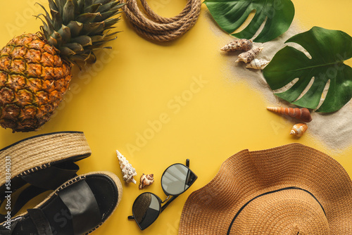 Colorful summer female fashion outfit flat lay. Straw hat, wicker shoes, pineapple over yellow background, top view, wide composition. Summer fashion, holiday concept