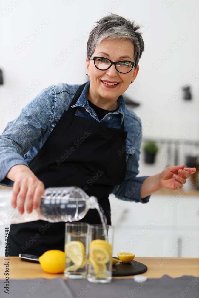 Happy senior woman in home interior pours water into glasses with lemon fresh homemade summer lemonade.