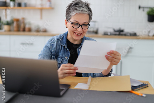 Slika na platnu Happy mature woman watching mail bank notice, sitting at kitchen table at home