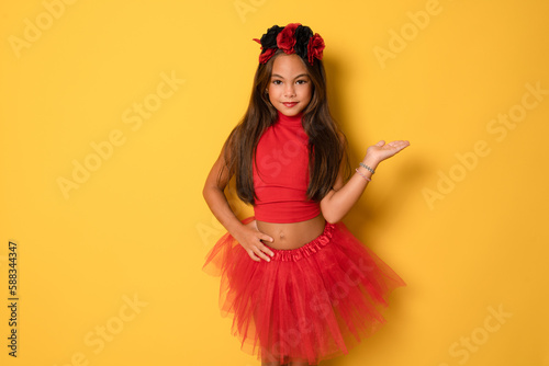 Cute smiling little kid girl in red halloween costume showing product with hand over yellow background. Halloween concept.
